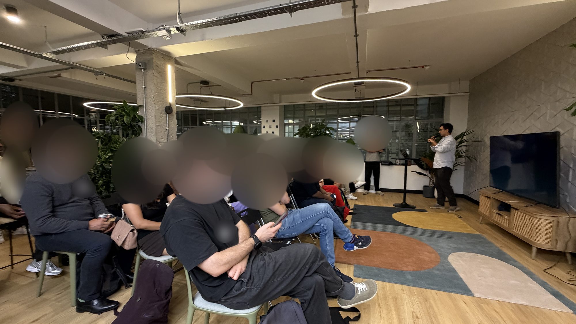 A room of seated people, their faces blurred, listening to Bilal Khizar speak at the front beside a large screen.
