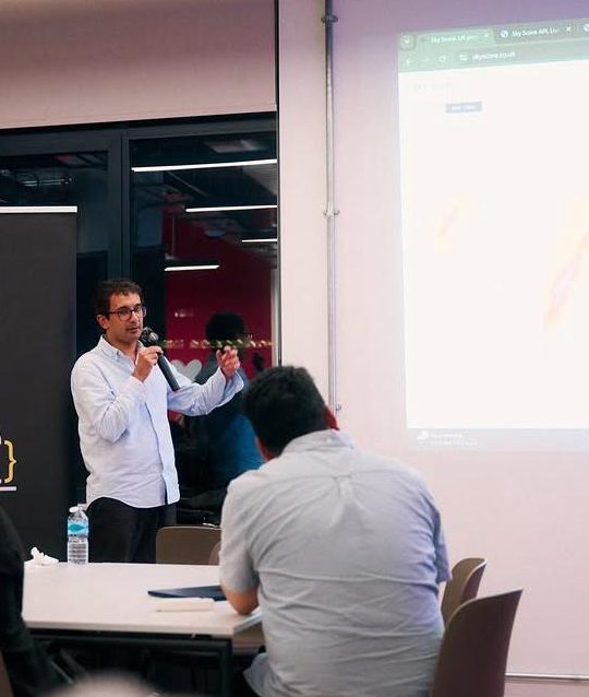 Bilal Khizar speaking with a microphone beside a projected Sky Score page.